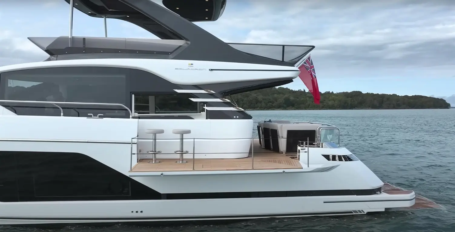 Modern yacht with sleek design, featuring outdoor seating, bar stools, and British flag on the rear in a serene water setting.